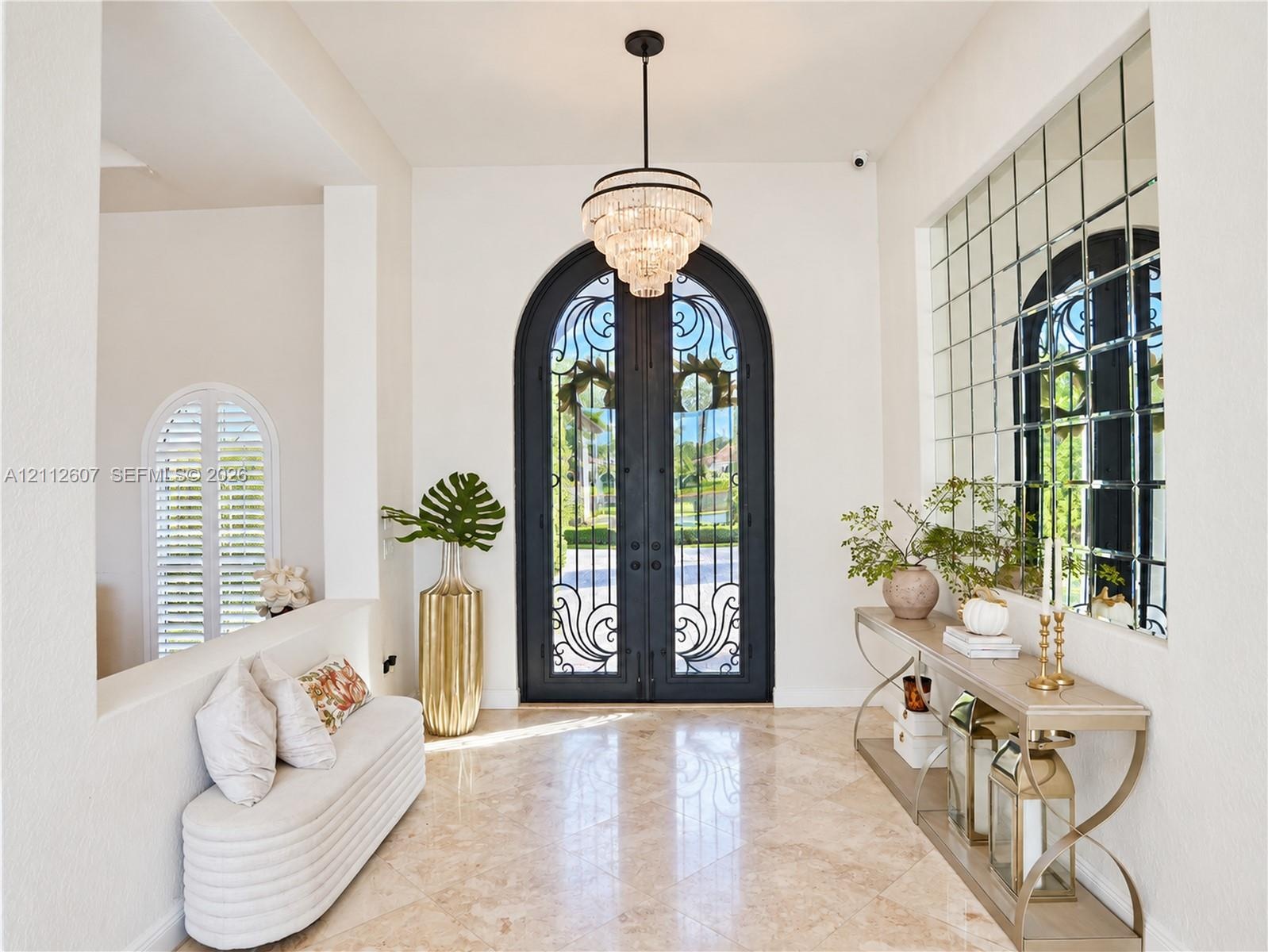 Foyer featuring arched double iron entry doors, light-toned stone flooring, a multi-tiered crystal chandelier, and an expansive grid-pattern wall mirr