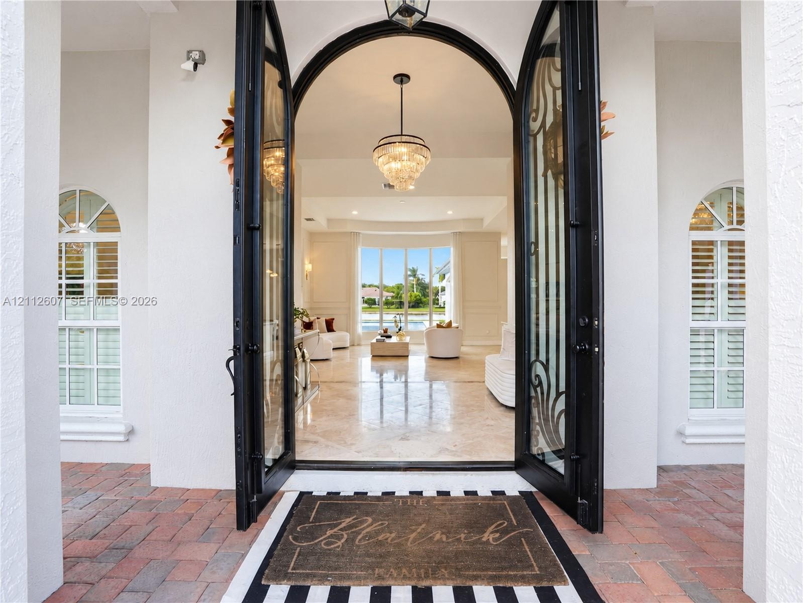 Arched double entry doors with decorative iron detailing, flanked by arched windows with plantation shutters, polished stone flooring, and a multi-tie