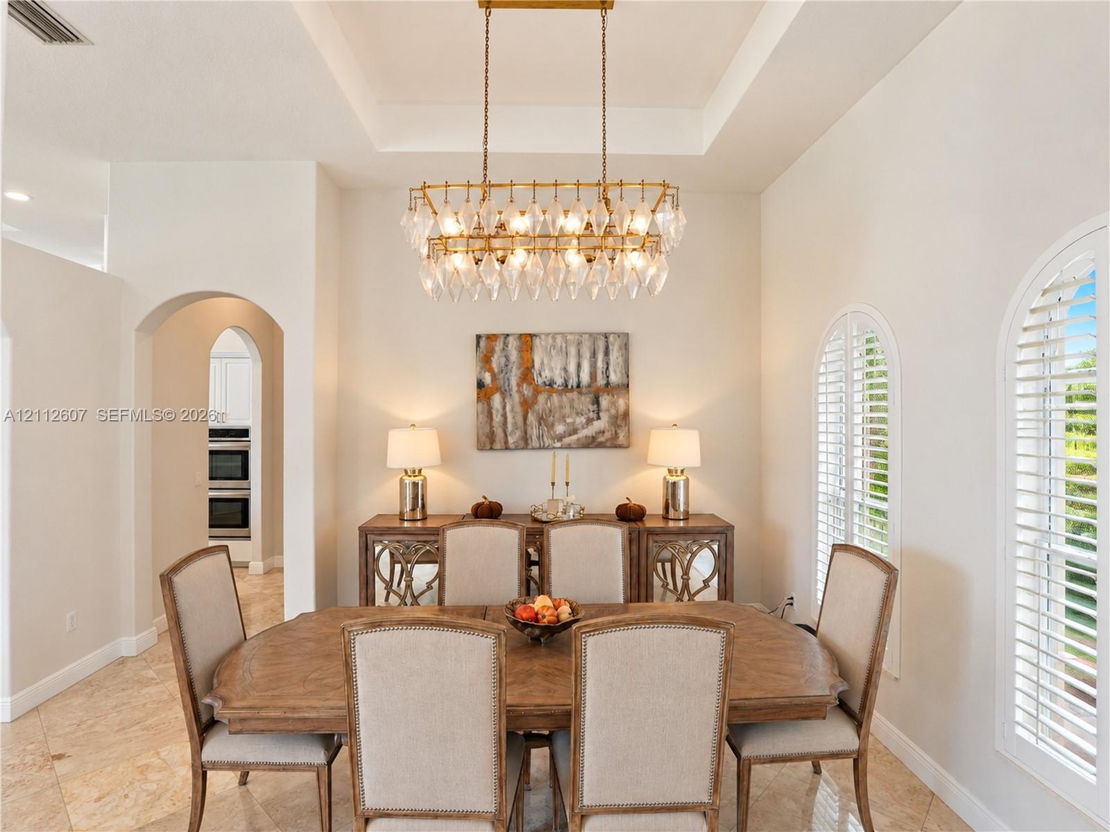 Tray ceiling with recessed lighting, tiered crystal chandelier, tile flooring, and arched windows featuring plantation shutters