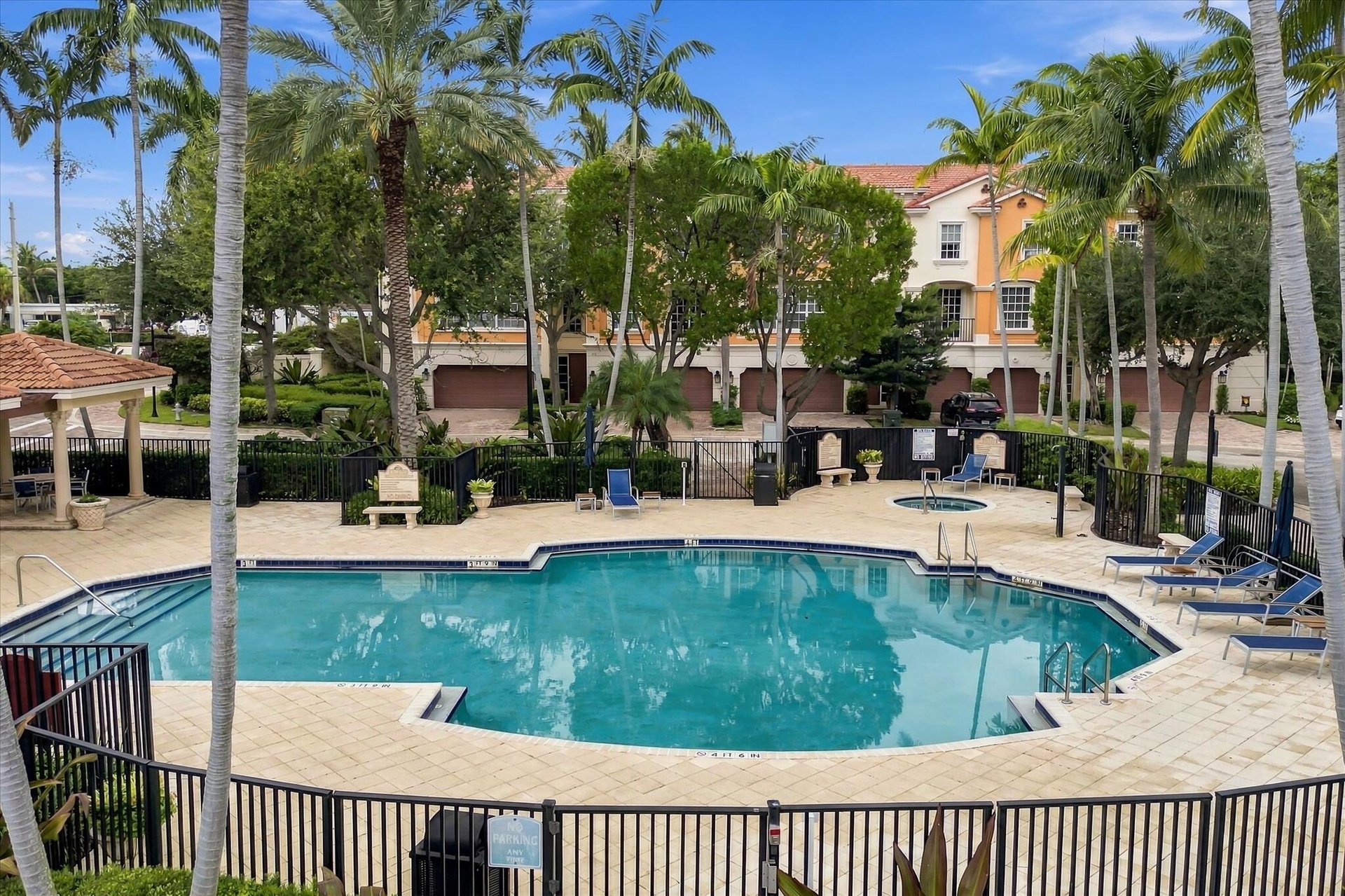 Swimming pool area featuring stone paver decking, a spa, covered pavilion, and metal perimeter fencing surrounded by mature palm trees