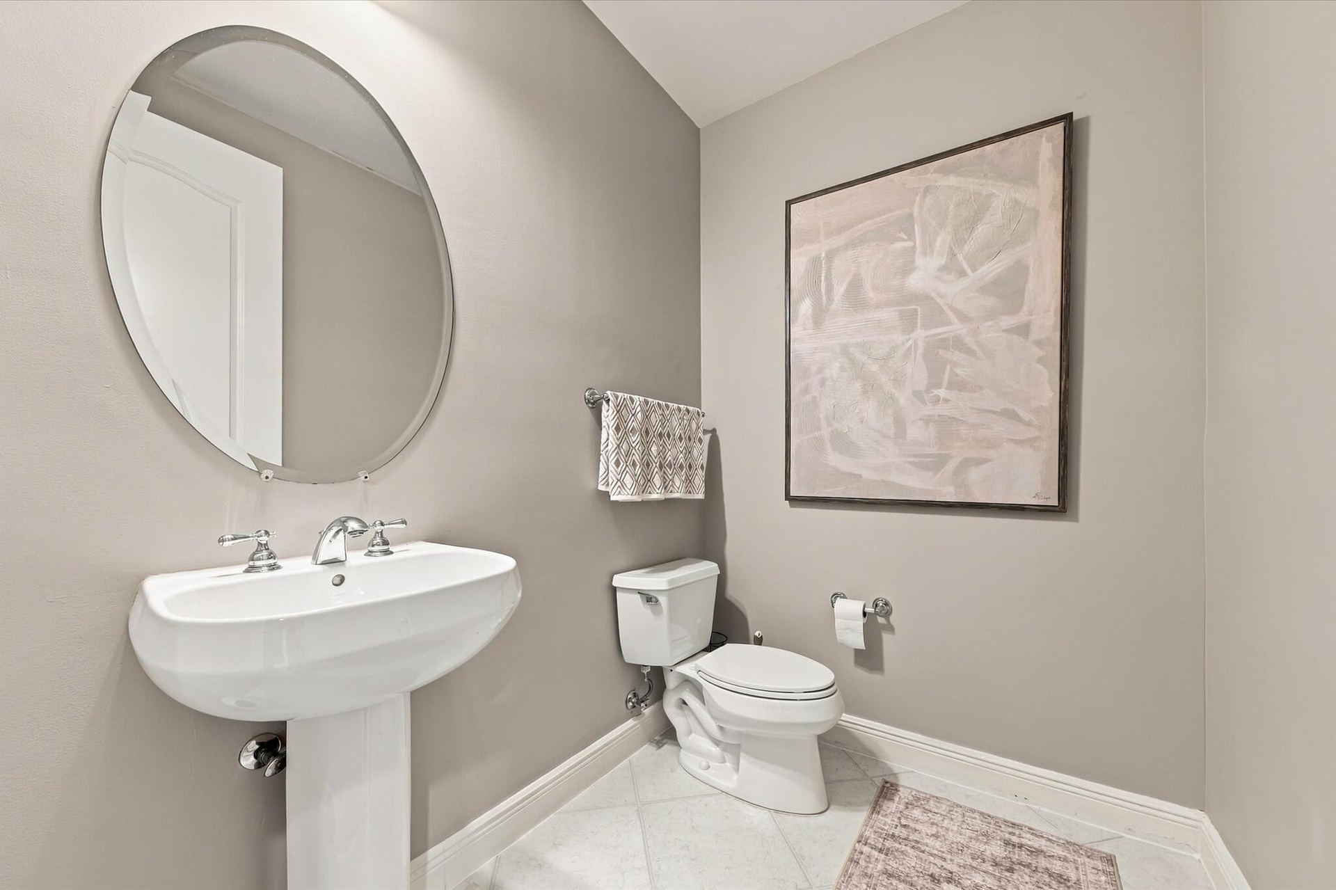 Oval mirror with a metallic frame, white porcelain pedestal sink, polished chrome widespread faucet, and ceramic tile flooring