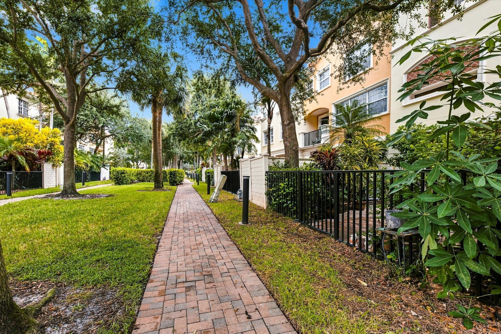 Brick paver walkway lined with mature trees, established landscaping, and black metal fencing