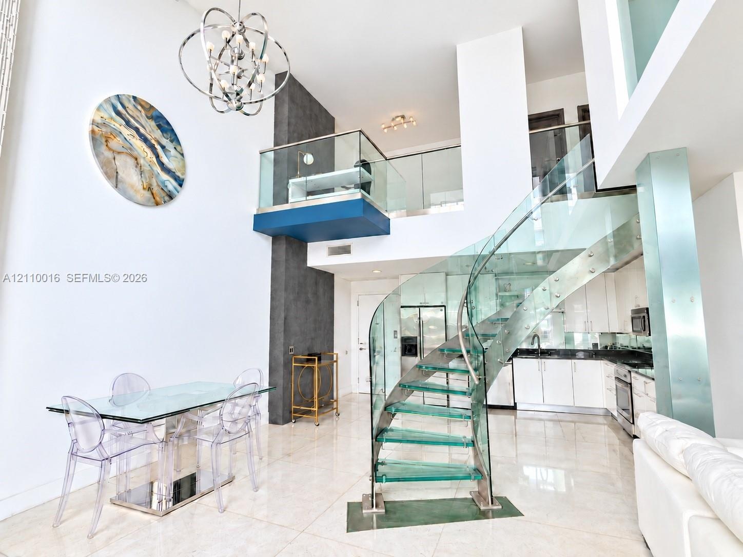Curved staircase featuring glass treads and stainless steel railings, two-story ceiling, polished stone flooring, custom metal chandelier, and a glass