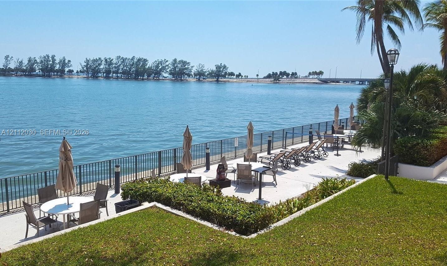 Waterfront patio featuring stone pavers, black metal perimeter fencing, manicured lawn, and mature landscaping