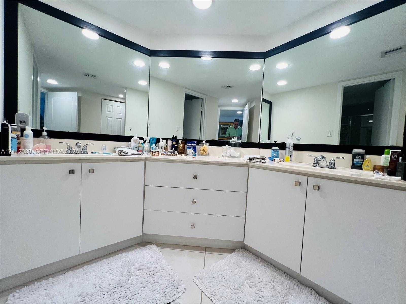 L-shaped dual vanity featuring white cabinetry, polished metal hardware, expansive wall-to-wall mirrors, and dual chrome faucets
