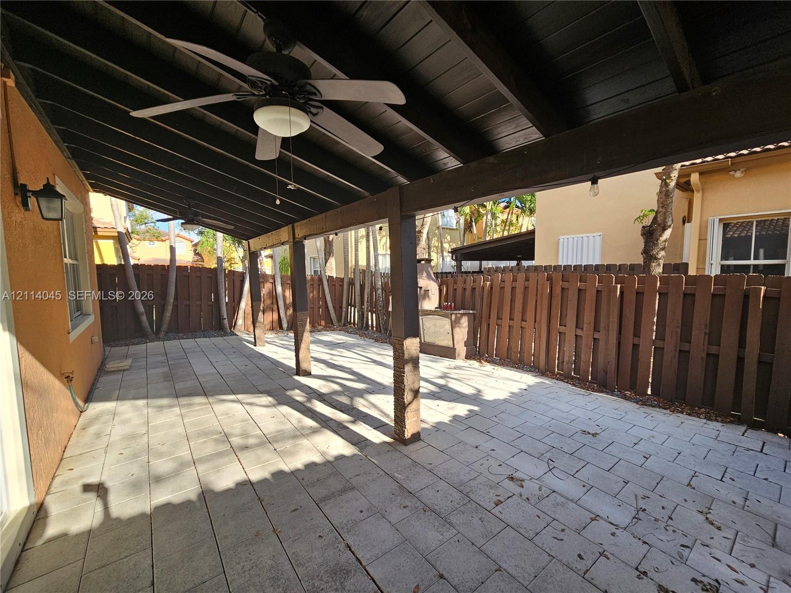 Covered patio featuring concrete paver flooring, dark-stained wood support beams, built-in ceiling fans, an exterior wall-mounted light fixture, and a
