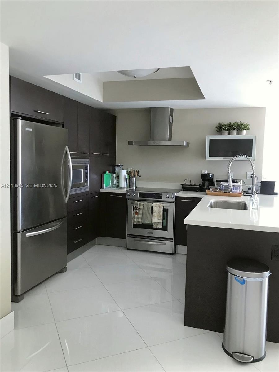 Kitchen featuring dark wood cabinetry, white stone-surface countertops, stainless steel appliances, a stainless steel chimney-style range hood, and po