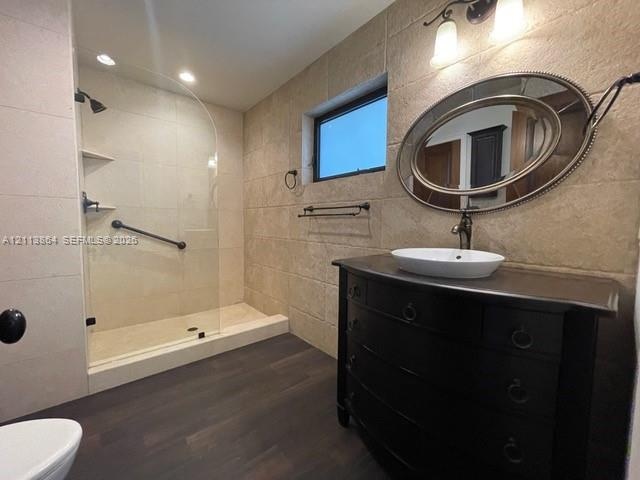 Bathroom featuring wood-finish flooring, textured stone-style wall tile, a glass-enclosed walk-in shower with integrated shelving, and a dark-toned va