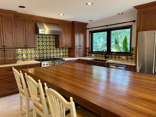 Kitchen featuring custom wood cabinetry, a large wood-topped center island, patterned tile backsplash, stainless steel range with a matching vent hood