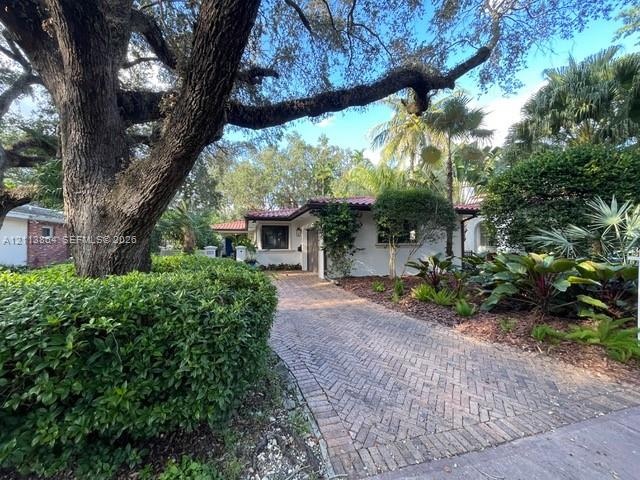 Single-story exterior featuring a red tile roof, herringbone-pattern brick driveway, mature canopy trees, and established tropical landscaping