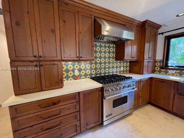 Custom wood cabinetry, stainless steel professional-grade gas range, stainless steel vent hood, patterned tile backsplash, and light-toned solid surfa