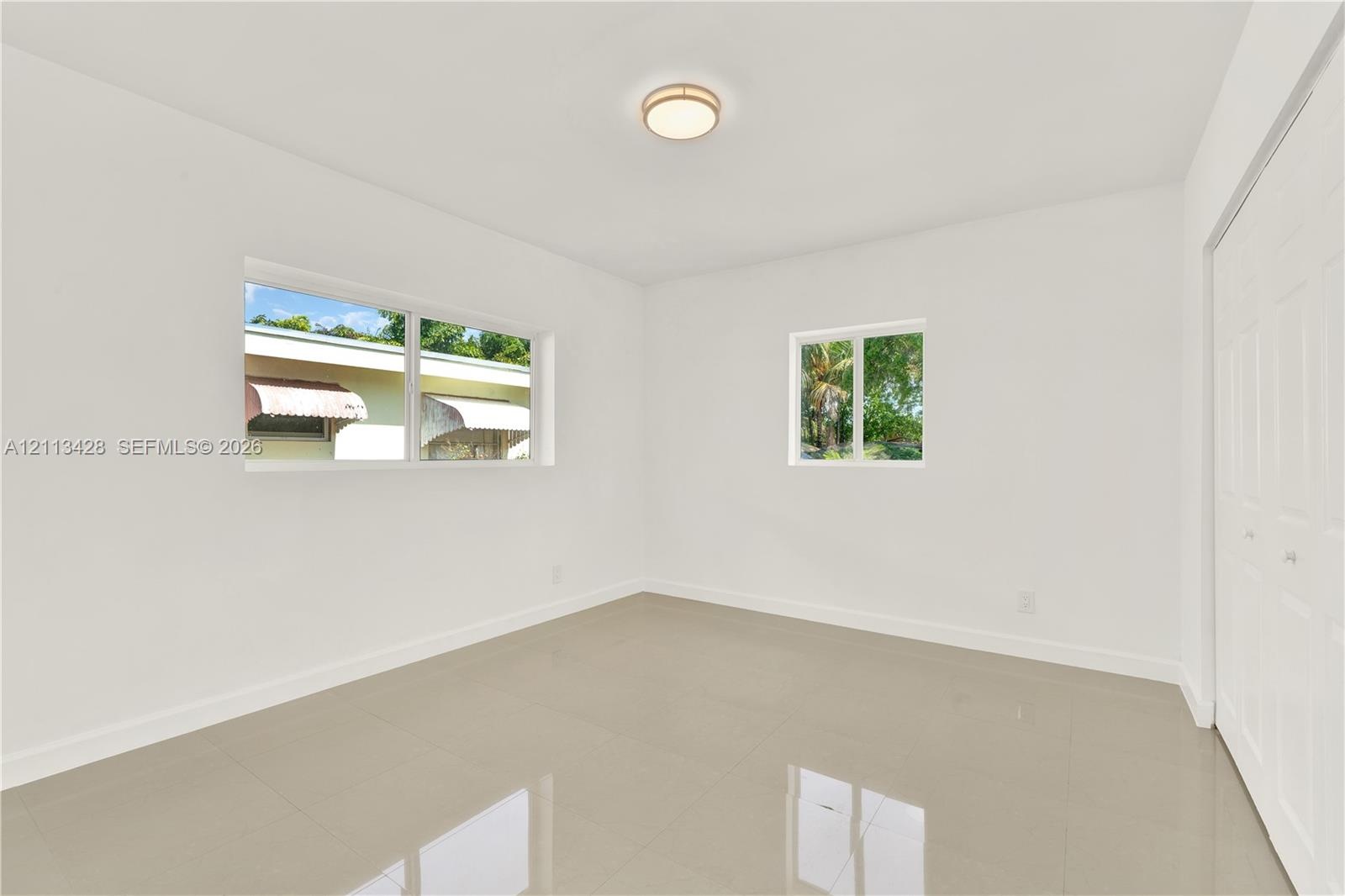 Neutral-toned tile flooring, dual windows, minimalist ceiling-mounted light fixture, and floor-to-ceiling closet with paneled doors