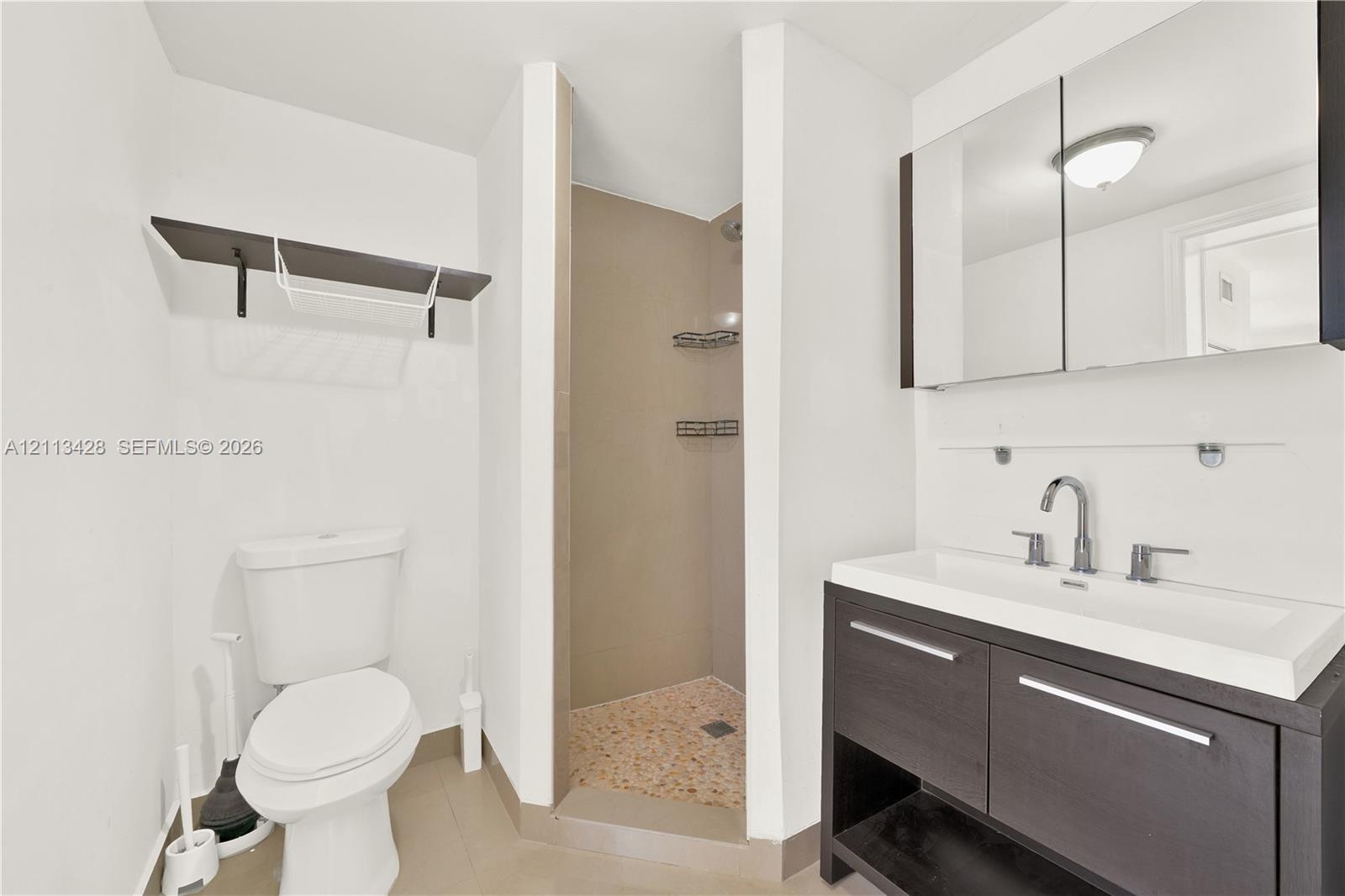 Bathroom featuring a vanity with dark cabinetry, a rectangular sink with a chrome faucet, a mirrored medicine cabinet, a walk-in shower with pebble-st