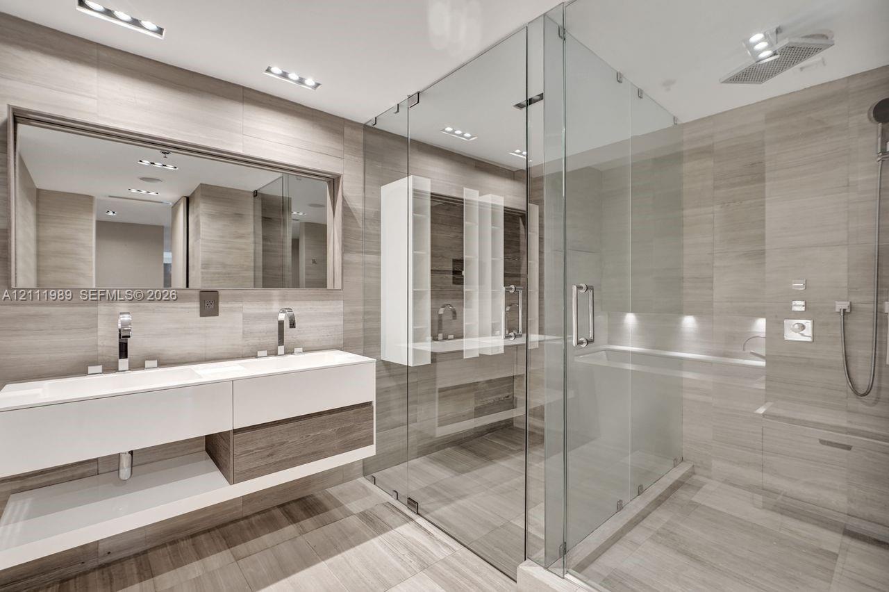 Floating double vanity with integrated sinks, large wall-mounted mirror, glass-enclosed walk-in shower with overhead rainfall fixture, and floor-to-ce