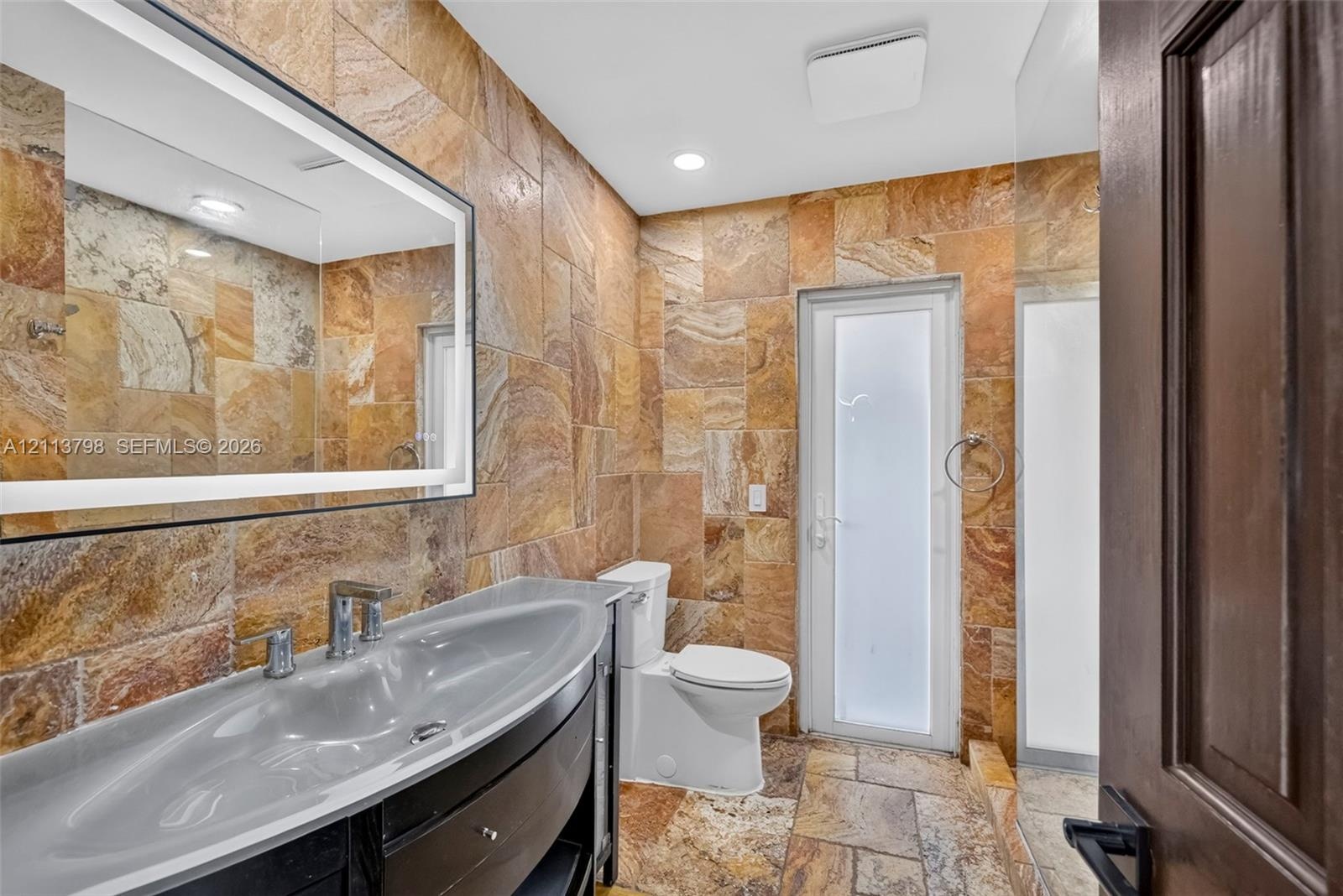 Stone tile wall and floor surfaces, custom vanity with integrated basin, LED-backlit mirror, and frosted glass paneled door