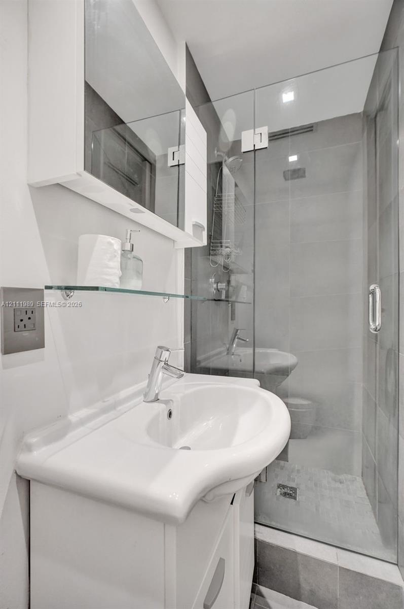 Wall-mounted medicine cabinet with mirror, pedestal-style vanity, glass-enclosed shower, wall-mounted glass shelving, and recessed ceiling lighting