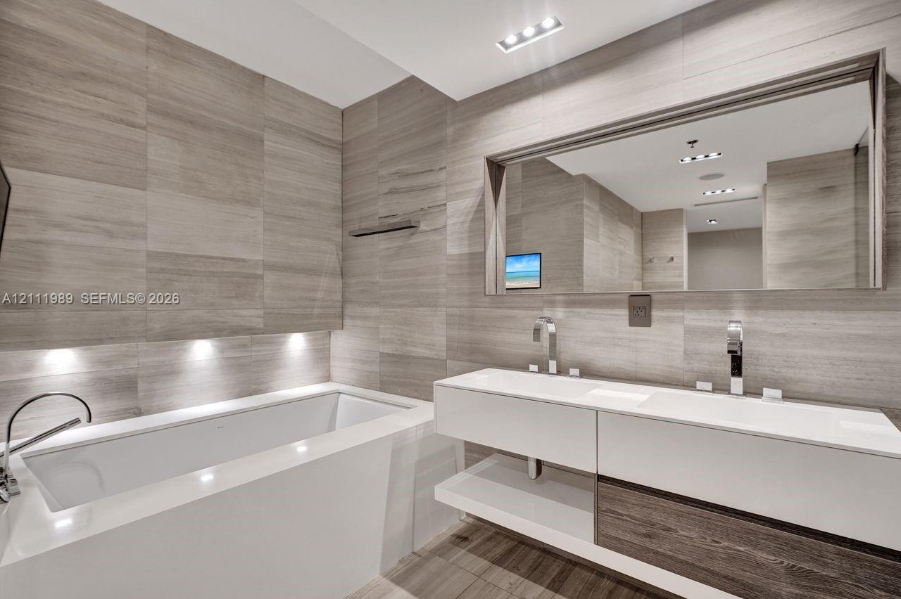 Modern floating dual-vanity with integrated sinks, floor-to-ceiling stone wall tiling, built-in soaking tub, and wall-mounted faucets