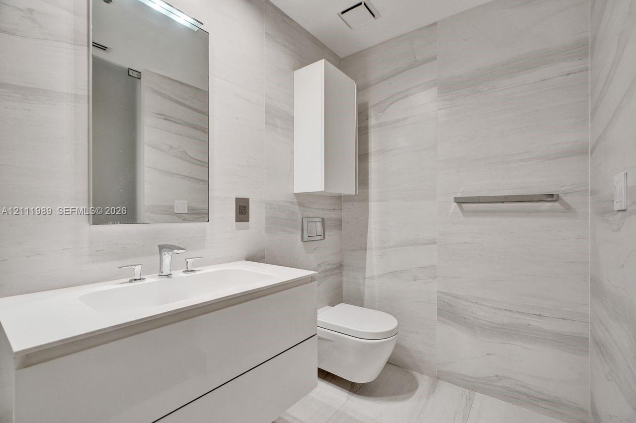 Floating vanity with a white countertop and integrated sink, wall-mounted toilet, floor-to-ceiling stone-style wall tile, frameless mirror, and wall-m