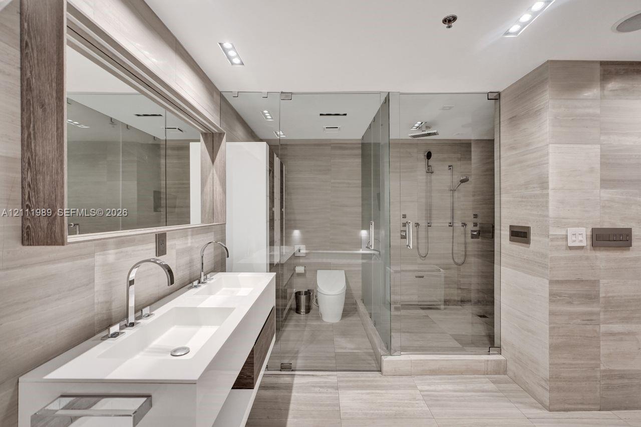 Floating dual-sink vanity with a solid-surface countertop, full-width mirror with wood-finish frame, frameless glass-enclosed shower, and stone-tiled 