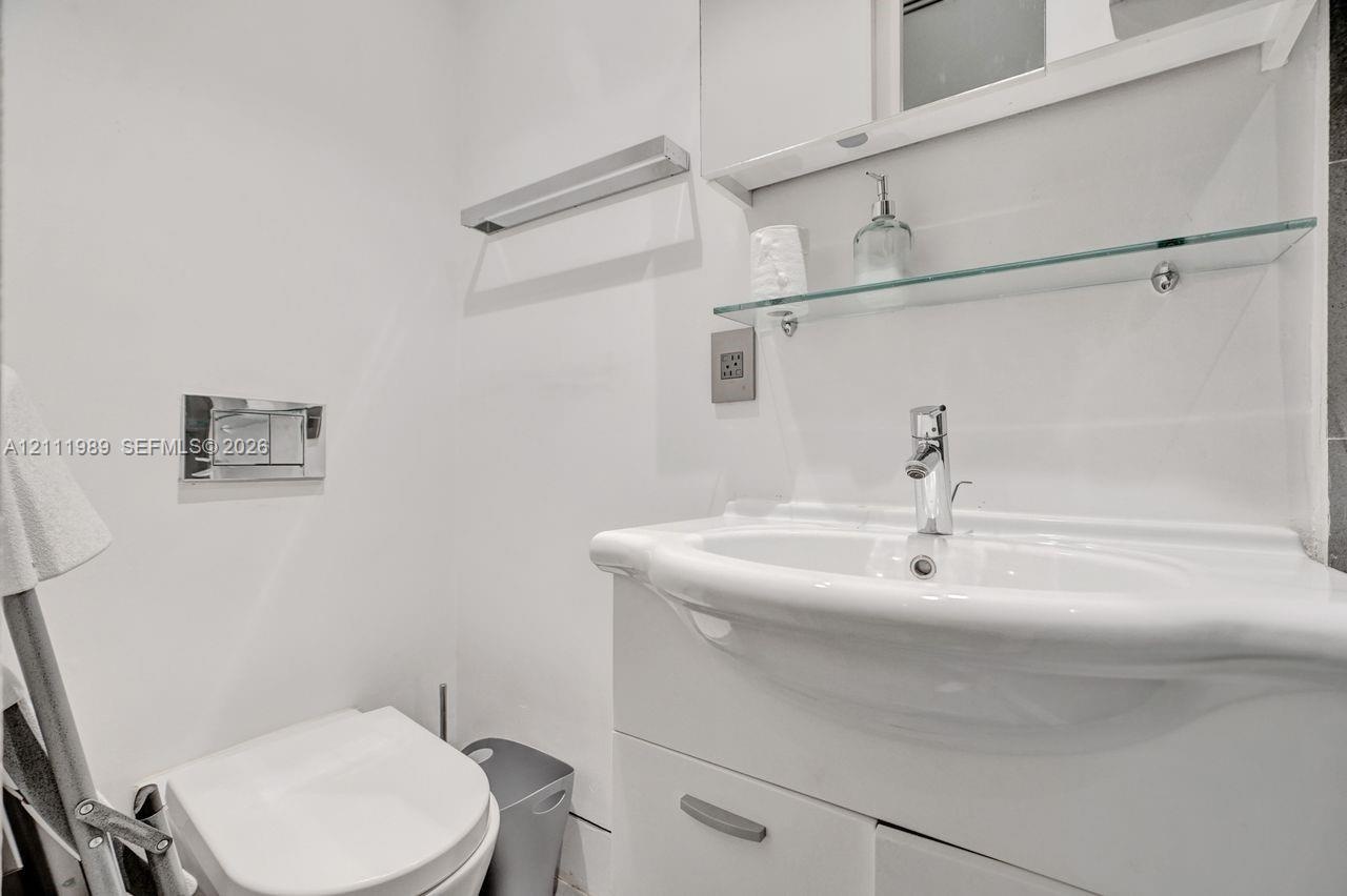 Wall-mounted toilet with chrome flush plate, white vanity cabinet, integrated porcelain sink with single-lever faucet, and glass shelving