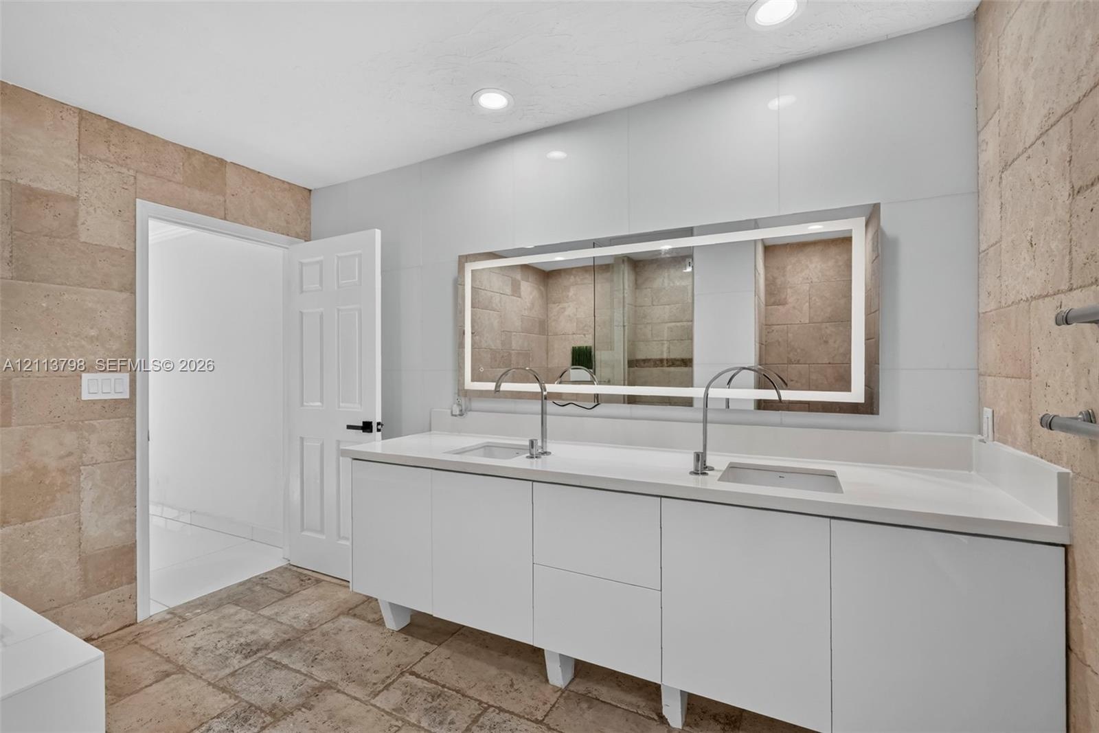 Double vanity with white cabinetry, solid surface countertop, rectangular undermount sinks, tall polished metal faucets, and a large backlit wall-moun