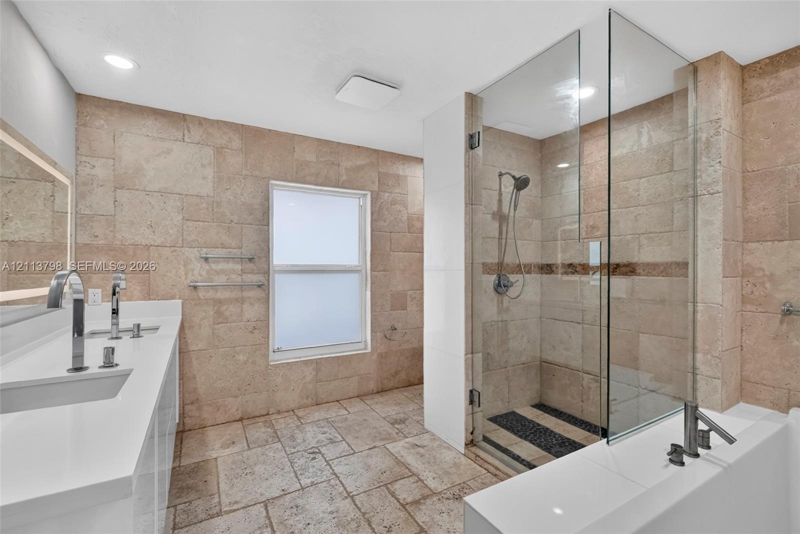 Bathroom featuring natural stone wall and floor tiling, double-basin vanity with solid-surface countertop, glass-enclosed walk-in shower with rainfall