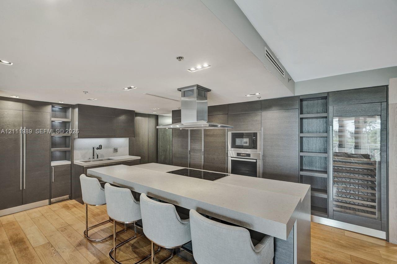 Kitchen featuring dark wood-finish cabinetry, a waterfall-edge center island with an integrated cooktop, a stainless steel chimney-style range hood, b