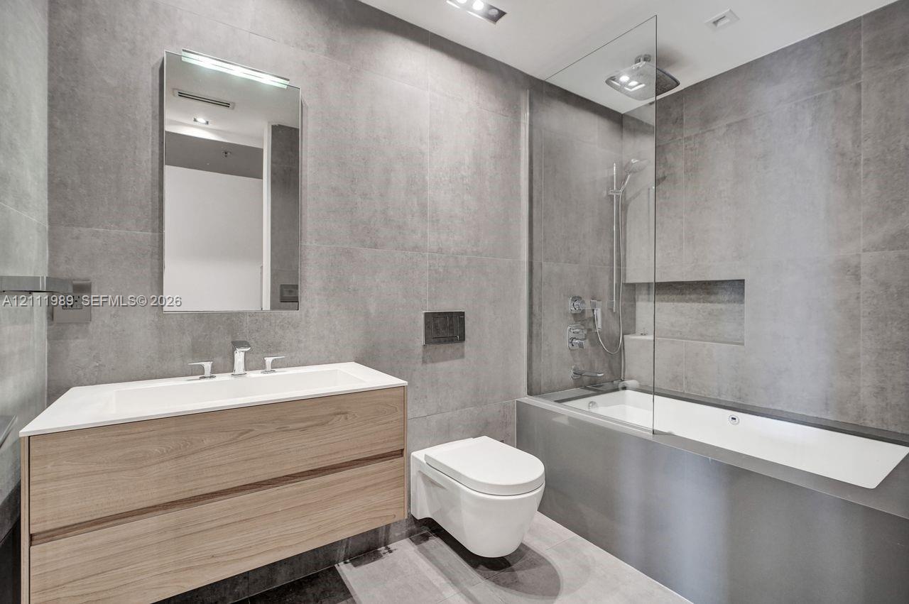 Bathroom featuring large-format grey tile wall cladding, wood-finish floating vanity with an integrated basin, wall-mounted toilet, and a glass-enclos