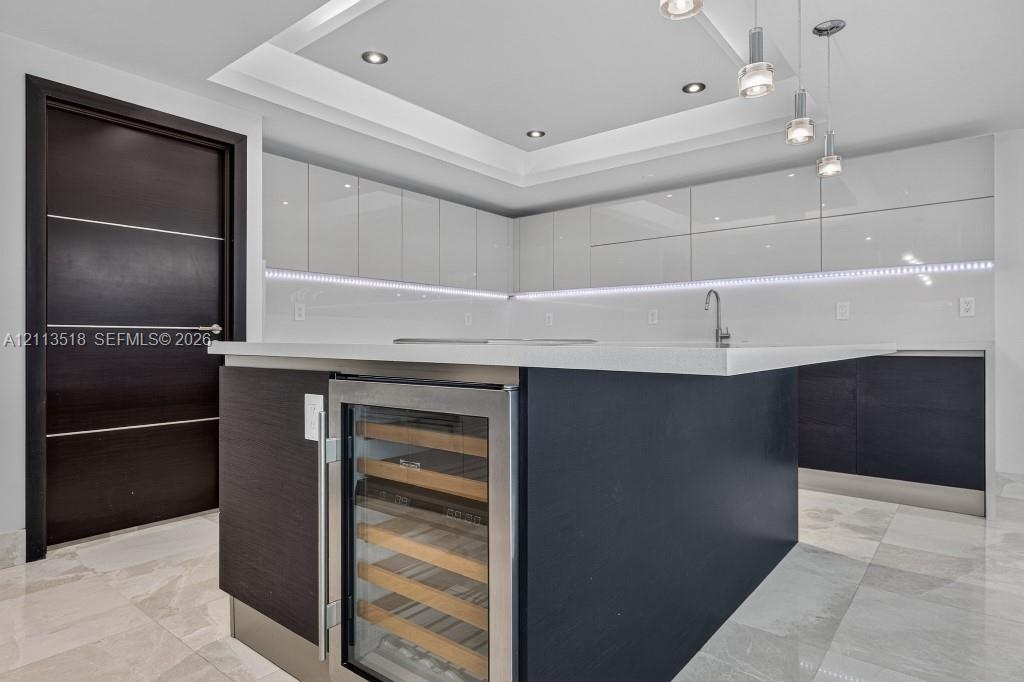 Kitchen featuring high-gloss cabinetry, a central island with a built-in wine refrigerator, stone countertops, recessed tray ceiling lighting, and til