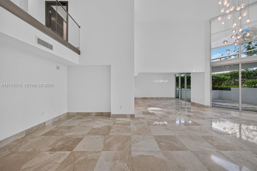 Double-height ceilings featuring floor-to-ceiling windows, polished stone flooring, a multi-pendant chandelier, and a glass-paneled mezzanine railing