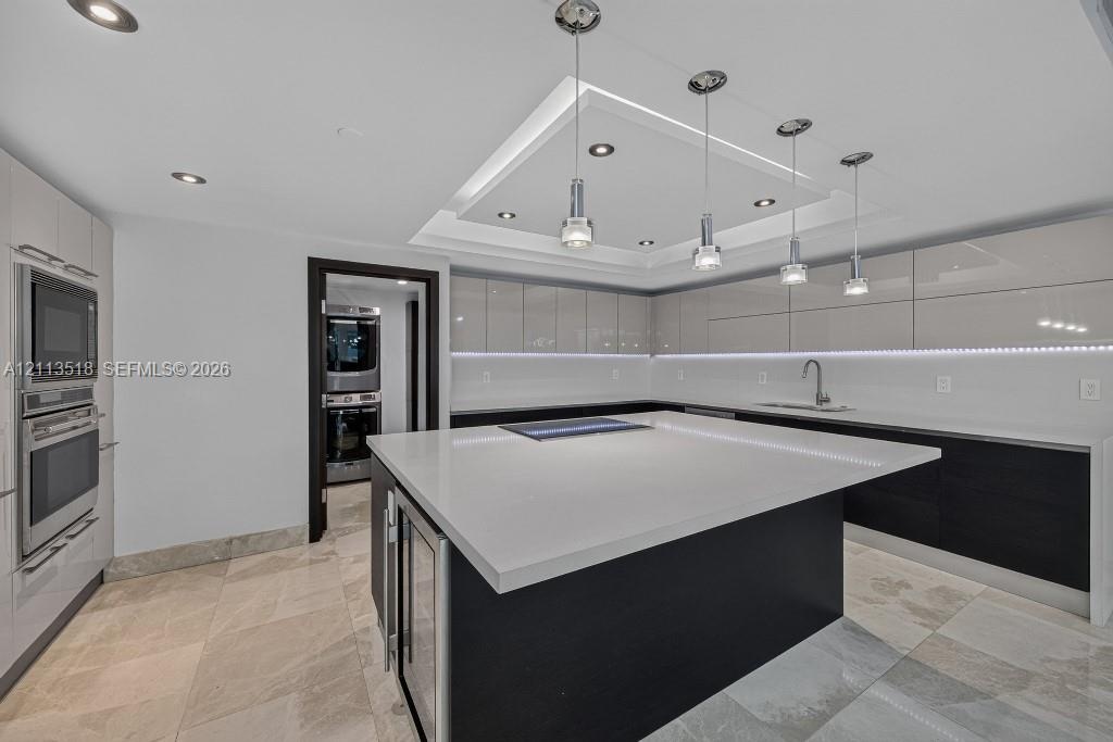Kitchen featuring high-gloss cabinetry, a central island with a solid-surface countertop, recessed tray ceiling lighting, stainless steel built-in app