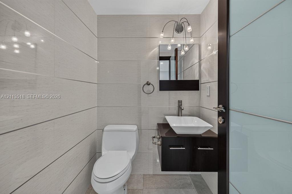 Bathroom featuring floor-to-ceiling large-format tile, wall-mounted dark wood vanity with a white vessel sink, multi-bulb wall-mounted lighting, and a