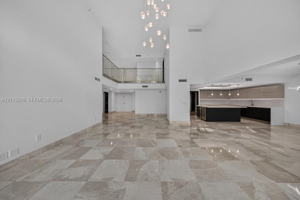 Expansive interior featuring high ceilings, stone flooring, a multi-tiered glass pendant chandelier, a glass-railed mezzanine, and kitchen cabinetry w