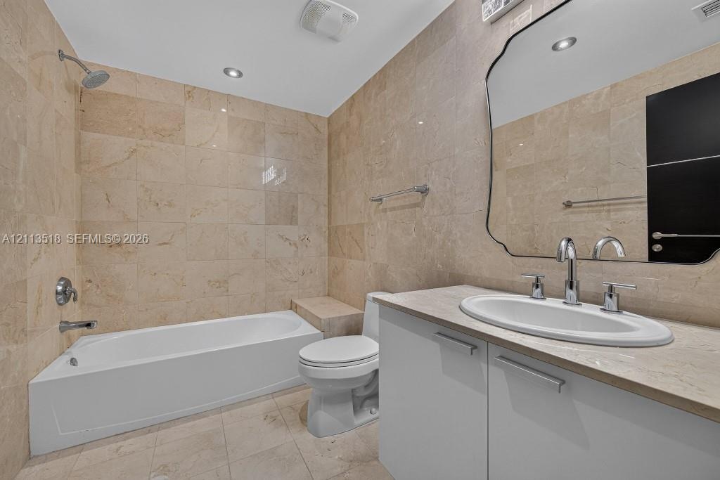 Full-height natural stone wall tiling, white vanity with an integrated countertop, oval undermount sink, decorative wall mirror, and built-in tub surr