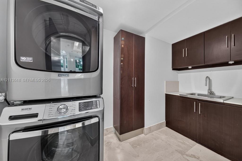 Laundry area featuring dark wood-finish cabinetry, a tall pantry unit, a stainless steel undermount sink with a gooseneck faucet, light-toned stone-fi