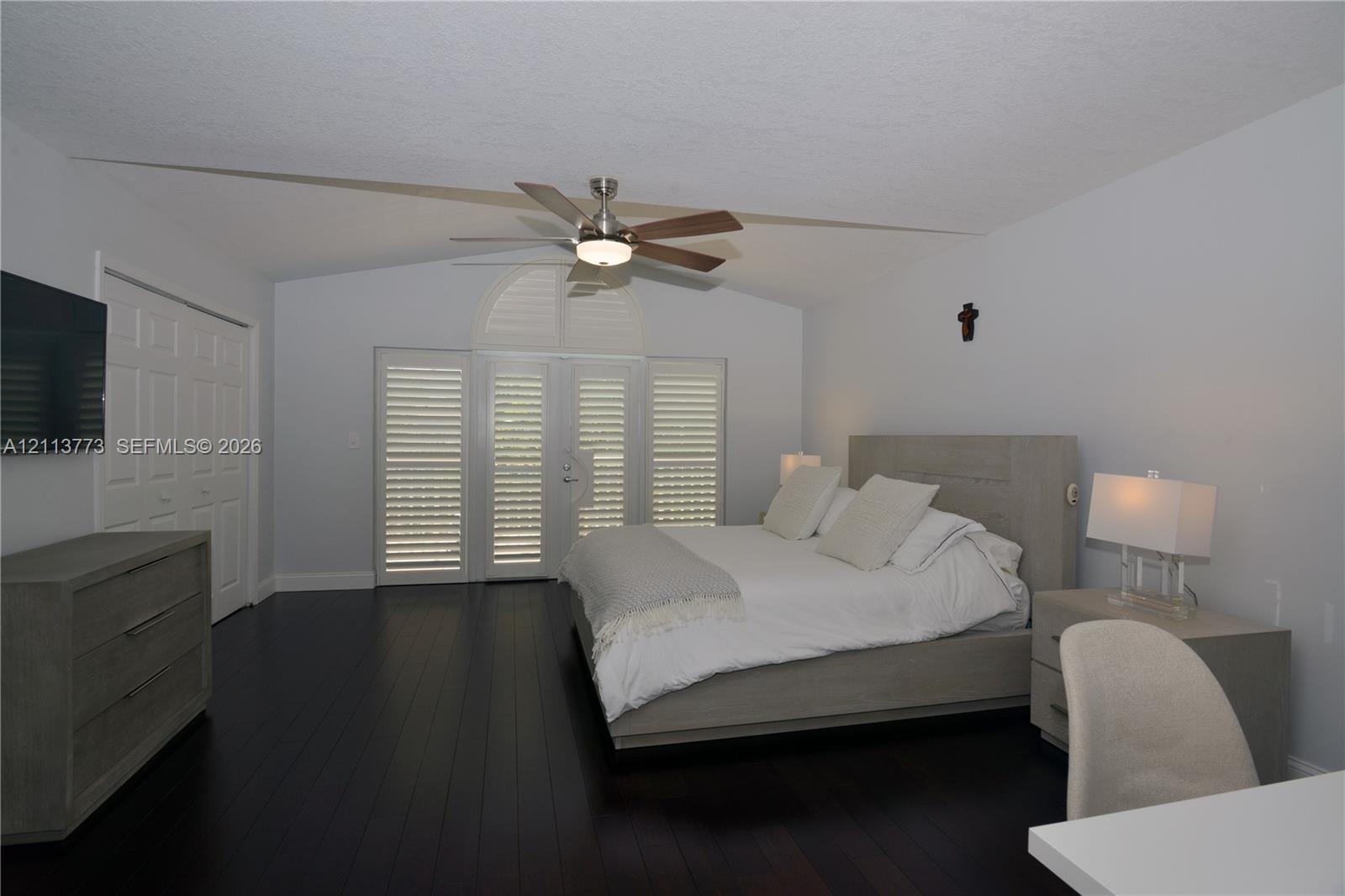 Vaulted ceilings with a central ceiling fan, dark wood-finish flooring, and white louvered plantation shutters on the double doors and windows