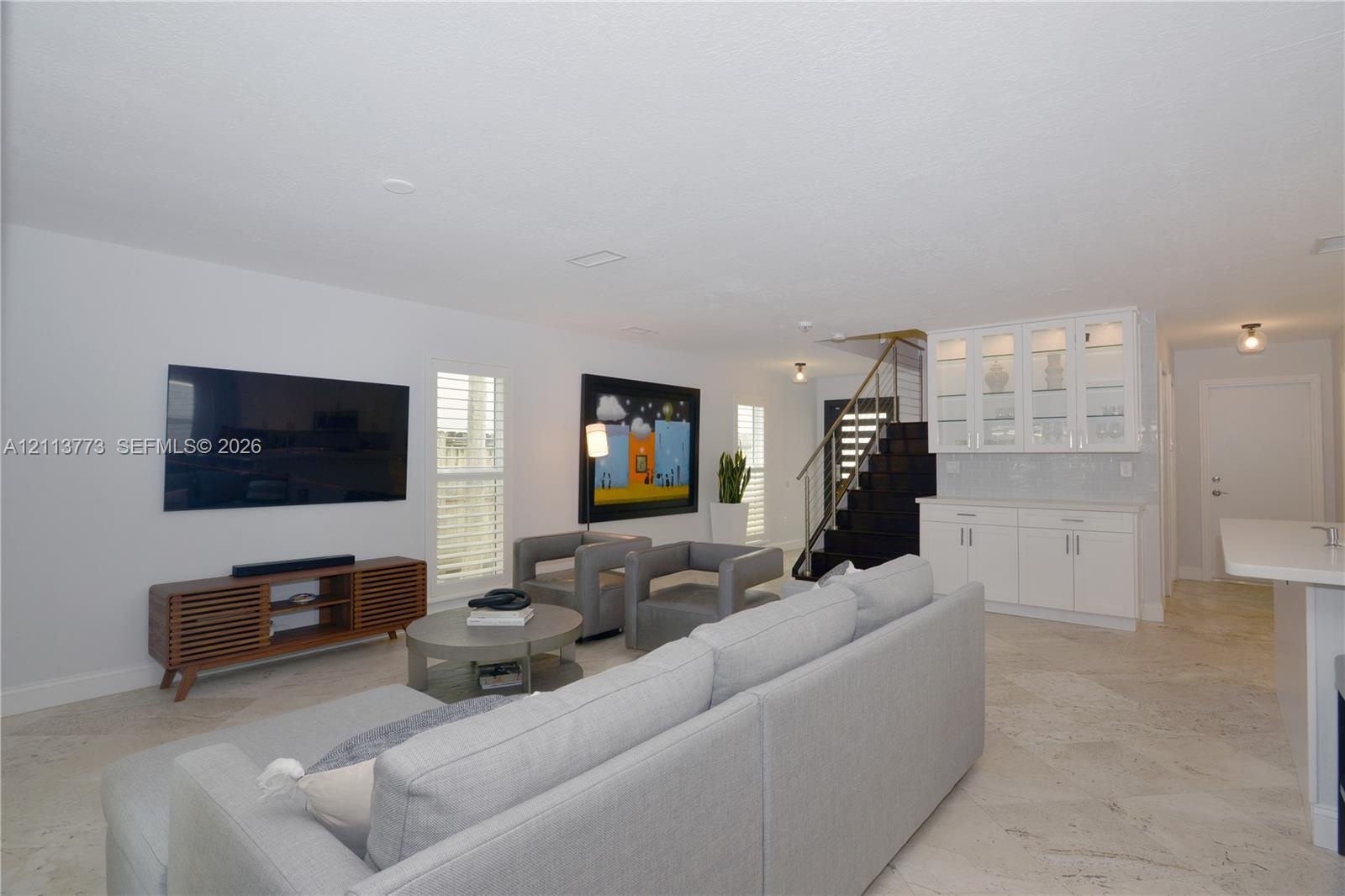 Open-concept interior featuring light stone-finish flooring, white built-in cabinetry with glass display uppers, and a custom staircase with metal rai