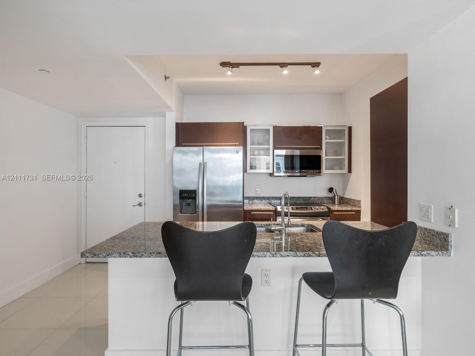 Kitchen featuring a stone countertop peninsula, stainless steel refrigerator and built-in microwave, flat-panel wood-finish cabinetry with glass-front