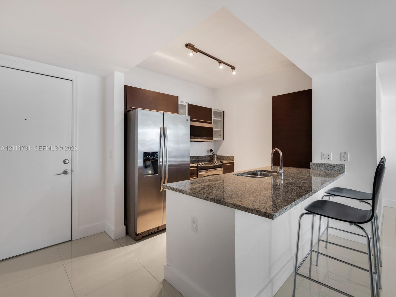 Kitchen featuring stone-surfaced countertops, a center island with an integrated sink, dark wood-finish cabinetry, stainless steel appliances, and tra