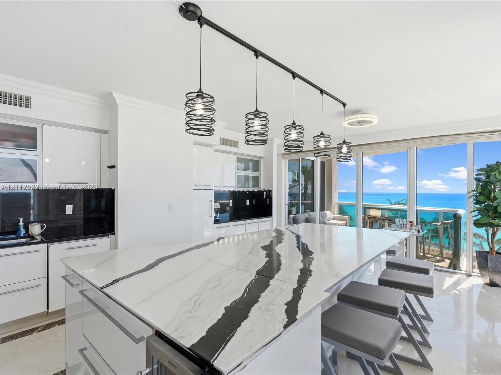 Kitchen featuring a large central island with veined stone countertop, high-gloss white cabinetry, black tile backsplash, and linear pendant lighting,