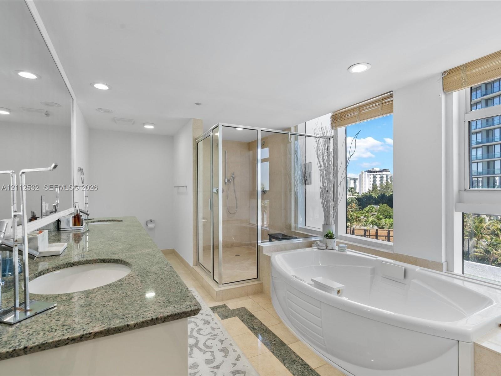 Dual-sink vanity with natural stone countertop, corner glass-enclosed shower, deep soaking tub, tile flooring with stone inlay, and large windows with