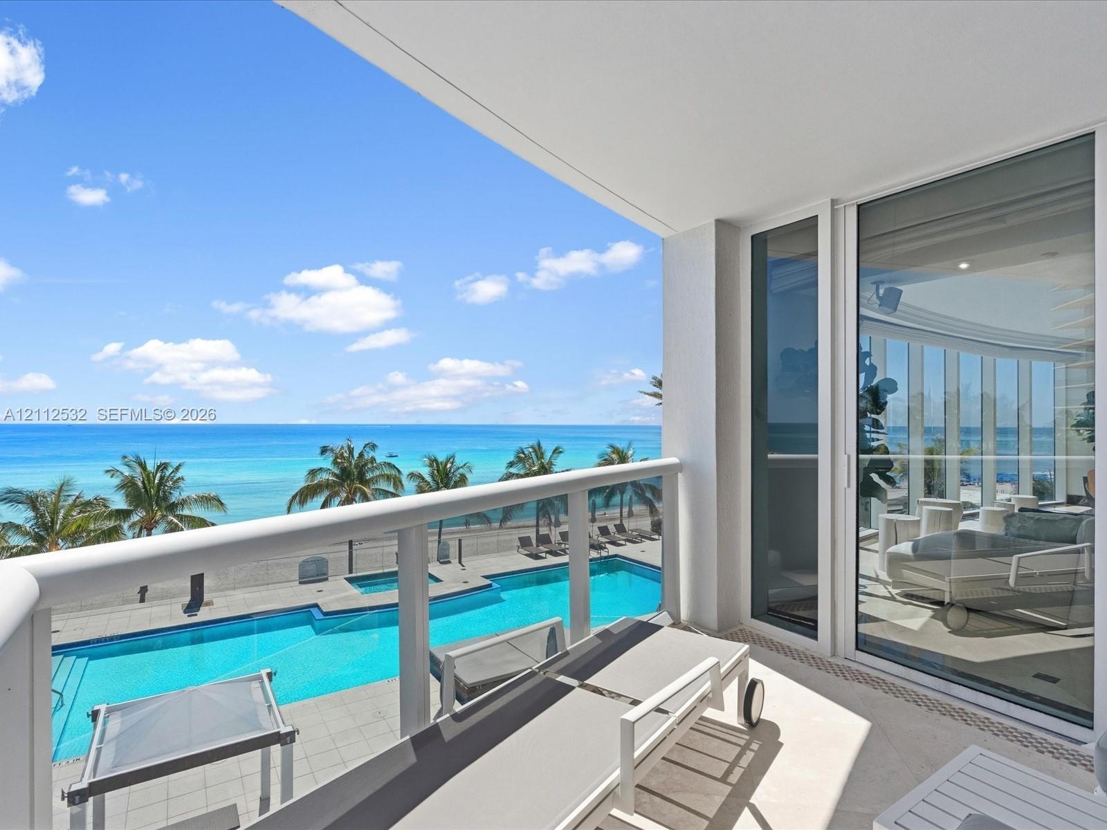 Covered balcony with glass and metal railings, overlooking a swimming pool and ocean backdrop, featuring floor-to-ceiling sliding glass doors