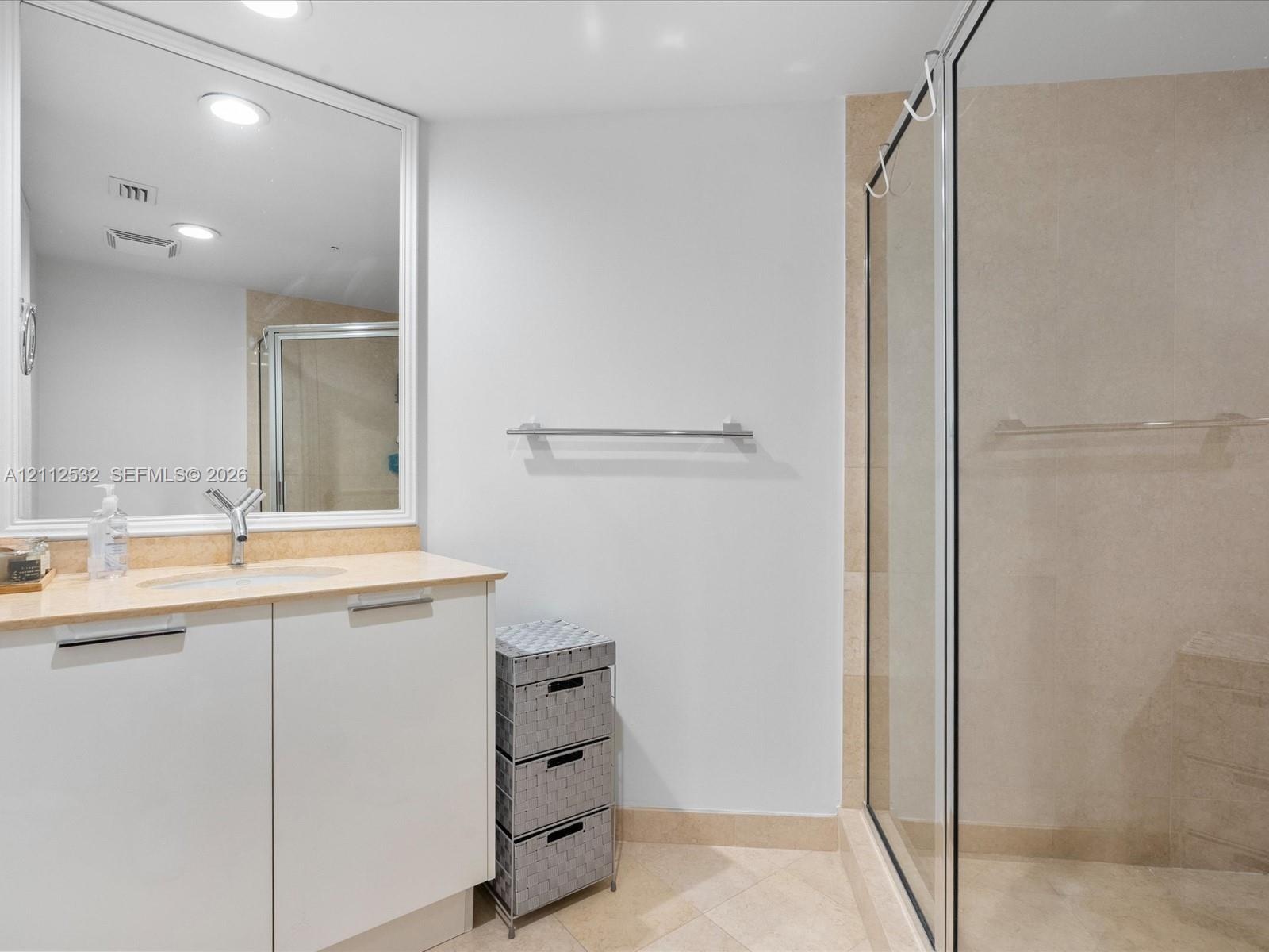 Bathroom vanity with a light-toned countertop, integrated sink, and white cabinetry, featuring a large framed mirror, stone-tile flooring, and a walk-