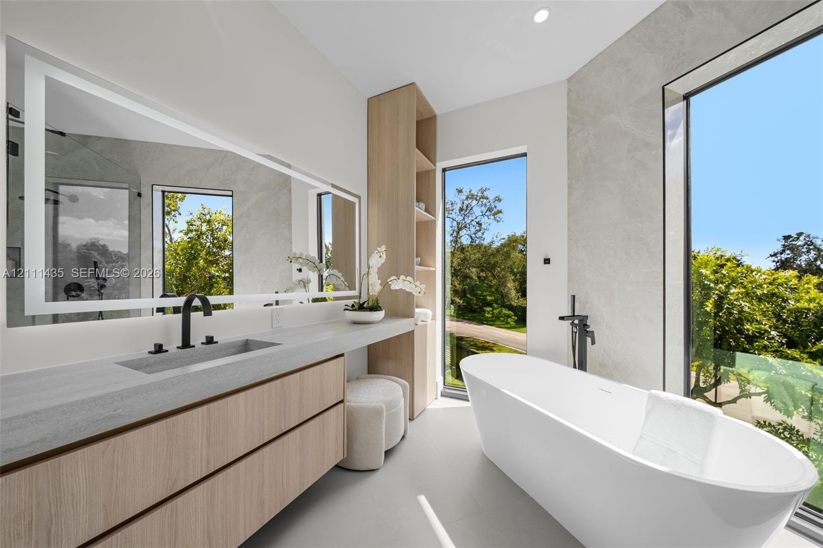 Floating wood-finish vanity with integrated sink, expansive LED-backlit mirror, freestanding soaking tub with floor-mounted faucet, and floor-to-ceili