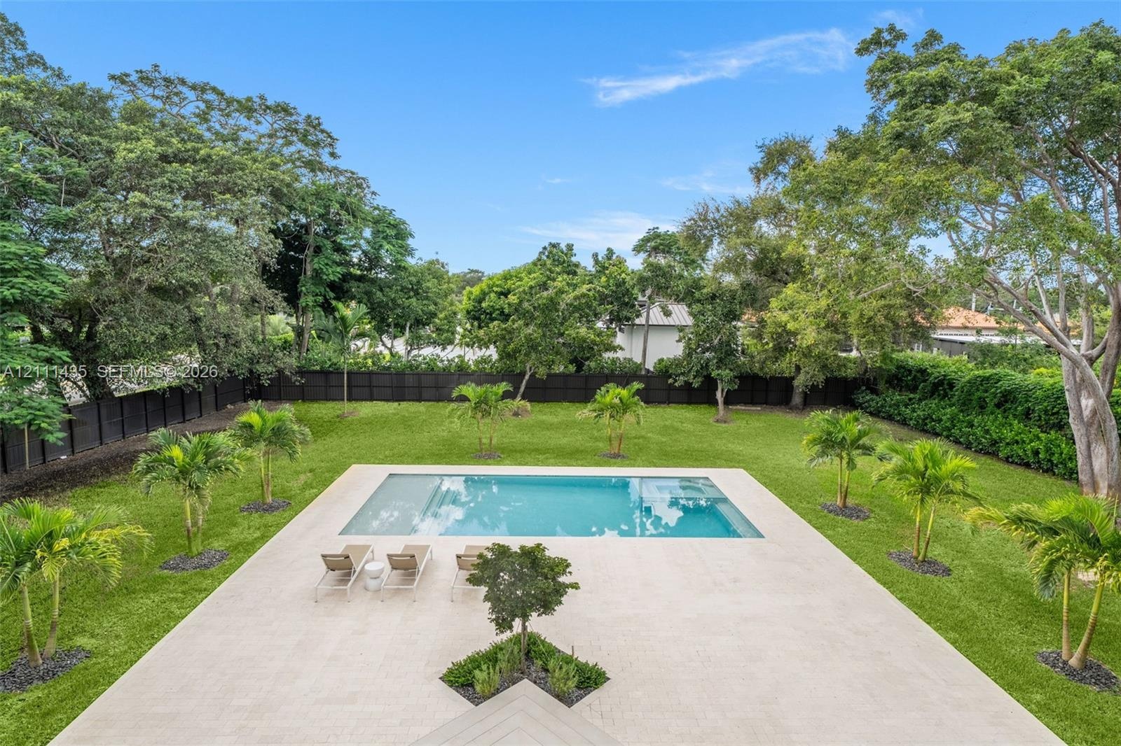 Expansive backyard featuring an in-ground swimming pool, light-toned concrete patio, manicured lawn, perimeter fencing, and mature trees