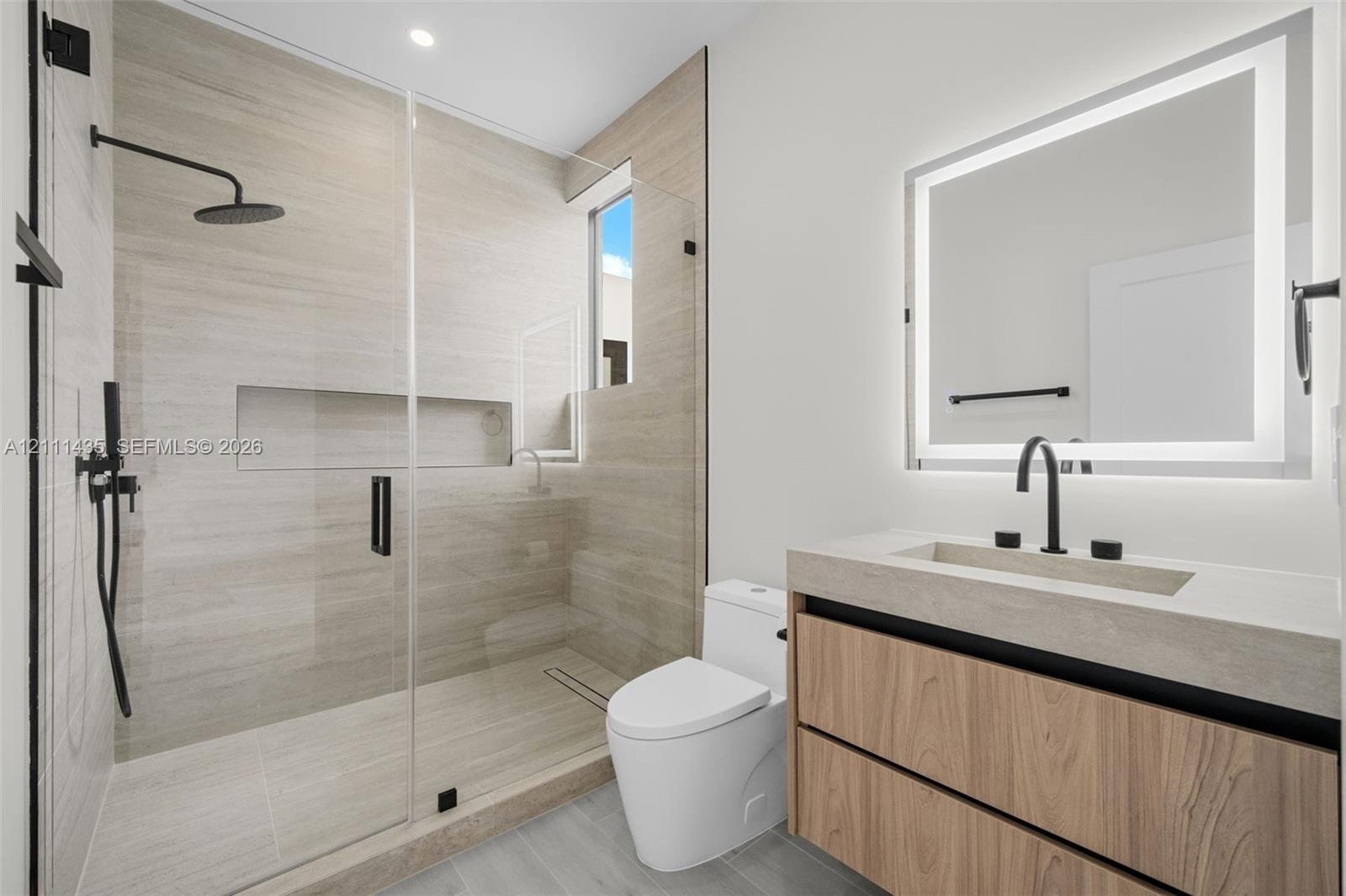 Bathroom featuring a floating wood-finish vanity, integrated stone-composite countertop, illuminated mirror, walk-in shower with floor-to-ceiling tile