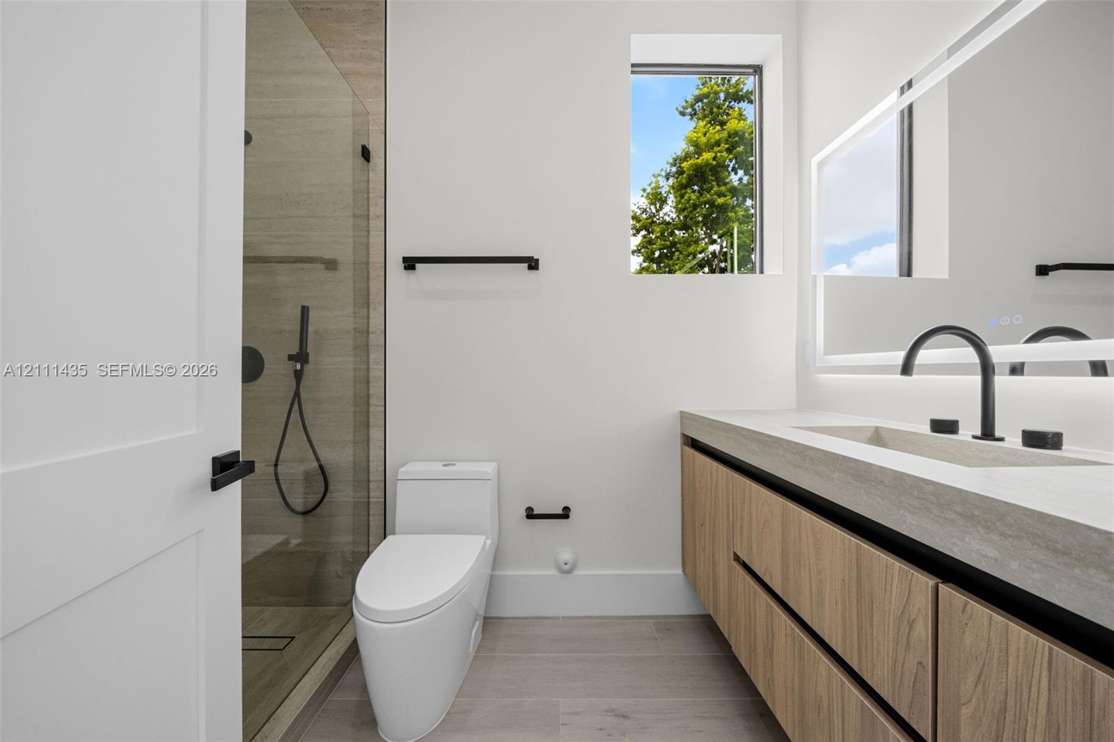 Floating vanity with wood-finish cabinetry, rectangular integrated sink, matte black hardware, glass-enclosed shower, and illuminated mirror