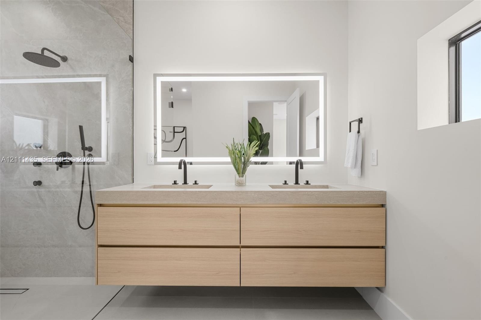 Floating wood-finish vanity with dual sinks, stone-surface countertop, LED-backlit mirror, and matte black fixtures, paired with a walk-in shower feat