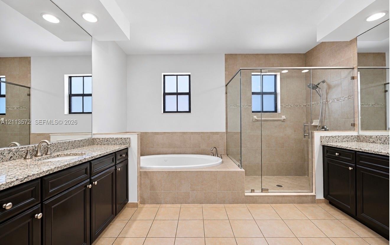 Dual vanities with stone countertops and dark cabinetry, large soaking tub with tile surround, and walk-in glass-enclosed shower with tiled walls