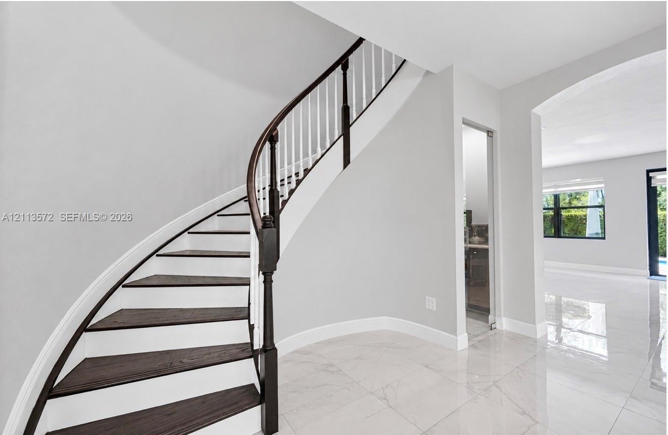 Curved staircase with dark-finished treads and white risers, decorative wood handrail, polished stone flooring, and arched entryway